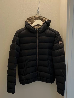 Colmar jacka - Snygg mörkblå pufferjacka från Colmar med huva och dragkedja framtill. Jackan har quiltad design, två snedställda fickor och Colmar-logga på ärmen. Fodret är ljust grått och materialet är polyester. Perfekt för kyliga dagar. Inga defekter passar xs/S