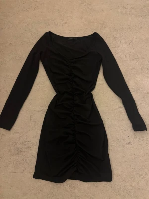 Svart långärmad omlottklänning - Svart figurnära klänning från Shein med lång ärm och snygg rynkning längs hela framsidan. Klänningen har en omlottliknande effekt och v-ringad hals, vilket ger en modern och stilren look. Perfekt för dig som gillar enkel men trendig stil.