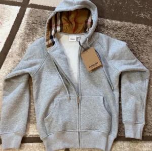 Grå hoodie från Burberry med dragkedja - Snygg grå hoodie från Burberry med klassiskt rutigt foder i huvan och silverfärgade detaljer på snörning och dragkedja. Tröjan har långa ärmar, två fickor fram och ribbade muddar. Perfekt för dig som gillar stilren design med lyxig touch.