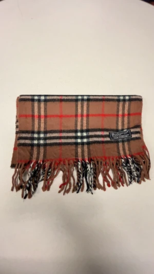 Rutig halsduk från Burberrys i ull - Snygg rutig halsduk från Burberrys i klassiskt beige, svart, vit och röd. Gjord av 100% lammull och har fransar i ändarna. Perfekt för att ge din outfit en lyxig touch och hålla dig varm under kyliga dagar.