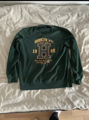 Grön college-tröja Brooklyn NYC - College-tröja i mörkgrönt med tryck i gult och blått framtill. Stor bokstav H och texten Brooklyn NYC 1985 ger en cool retro-vibe. Rund hals och långa ärmar, perfekt för chill dagar. Materialet är mjukt och känns som bomull.