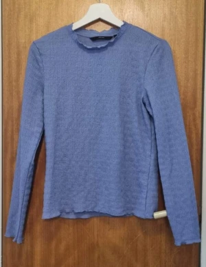 Blå långärmad tröja med volangkant - Blå långärmad tröja med volangdetaljer vid halsringning och nederkant. Materialet är mjukt och har en lätt struktur som ger en skön känsla. Perfekt att matcha med jeans eller kjol för en chill look.