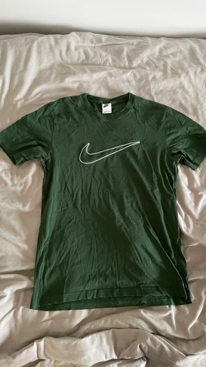 Grön Nike t-shirt med logga - Snygg grön t-shirt från Nike med den klassiska vita swoosh-loggan på bröstet. Modellen är kortärmad och har en rund halsringning. Perfekt för dig som gillar sportiga och stilrena plagg. Materialet är mjuk bomull som känns skönt mot huden.