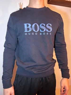 Mörkblå sweatshirt från Hugo Boss - Snygg mörkblå sweatshirt från Hugo Boss med stort BOSS-tryck i blått på bröstet. Tröjan har rund hals, långa ärmar och ribbade muddar vid ärmslut och nederkant. Perfekt för dig som gillar stilrena och klassiska märkesplagg. L som sitter som en M