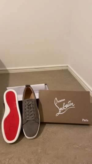 Christian Louboutin grå mocka sneakers - Säljer ett par gråa sneakers från Christian Louboutin i lyxig mocka med klassisk röd sula och vita kanter. Skorna har rund tå, snörning och diskret branding. Perfekta för dig som vill sticka ut med en stilren och exklusiv look.