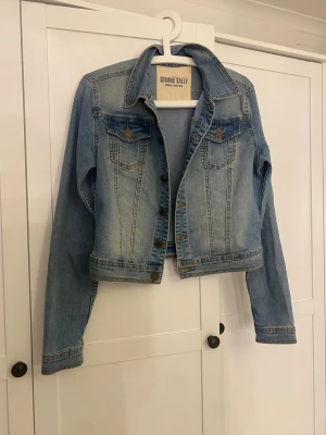 Ljusblå jeansjacka från Serious Sally - Helt ny Snygg ljusblå jeansjacka från Serious Sally med klassisk krage, bröstfickor med knappar och slitna detaljer. Jackan har långa ärmar och bronsfärgade knappar framtill. Perfekt för dig som gillar en chill och tidlös denimstil.