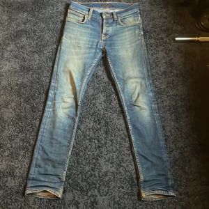 Blå slim jeans från Nudie Jeans - Snygga blå slim jeans från Nudie Jeans med slitningar och klassiska orange sömmar på bakfickorna. Jeansen har låg midja och smal passform, tillverkade i mjukt denimtyg. Perfekta för dig som gillar en smal siluett och coola detaljer. Notera att det är en lagning på insidan men inget som syns på utsidan, endast feta slitningar. Köpt för 1500 på NK.