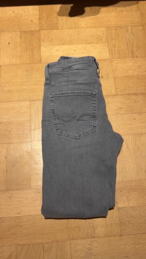Grå jeans från Jack & Jones - Snygga grå jeans från Jack & Jones med klassisk femficksdesign och raka ben. Jeansen har normal passform och är tillverkade i mjukt bomullsmaterial med lite stretch för extra komfort. Perfekt för dig som gillar stilrena och enkla jeans.