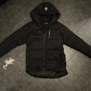 Sail Racing Jacka - Svart dunjacka från Sail Racing med huva och dragkedja framtill. Jackan har loggor på bröstet, ärmen och huvan samt snygga detaljer. Perfekt för kalla dagar och har en sportig vibe. Fylld med dun och har två fickor med dragkedja.