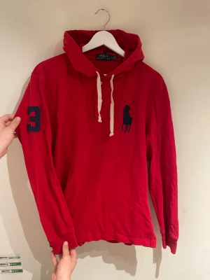 Röd hoodie från Polo Ralph Lauren - Snygg röd hoodie från Polo Ralph Lauren med stor marinblå logga på bröstet och en broderad siffra 3 på ärmen. Tröjan har huva med vita snören och långa ärmar. Perfekt för dig som gillar sportig och klassisk stil.  Storlek M passar S