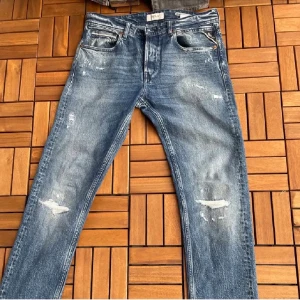 Replay Anbass blå jeans med slitningar - Säljer ett par Replay Anbass jeans i klassisk blå tvätt med snygga slitningar och distressed detaljer. Modellen har fem fickor, raka ben och normal passform. Jeansen är tillverkade i stretchigt denim för extra komfort och har Replay-logga på bakfickan.