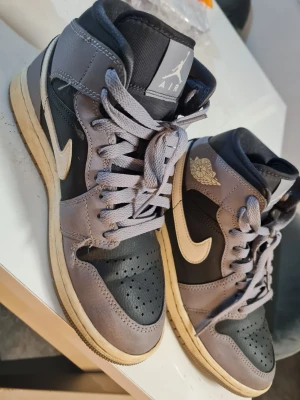 Nike Air Jordan 1 High svart/grå - Nike Air Jordan 1 High sneakers i svart och grått med beige detaljer. Klassisk high-top siluett med perforerad tå, snörning och ikonisk Air Jordan-logga på sidan. Ovandel i skinn och syntet, platt sula och rund tå. Snygga och tidlösa för dig som gillar streetstyle.