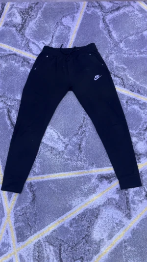 Svarta Nike mjukisbyxor med snörning - Svarta mjukisbyxor från Nike med klassisk logga på vänster lår. Byxorna har snörning i midjan, ribbade muddar vid bensluten och dragkedjeförsedda fickor. Skön passform och stilren look, perfekta för chill eller träning.