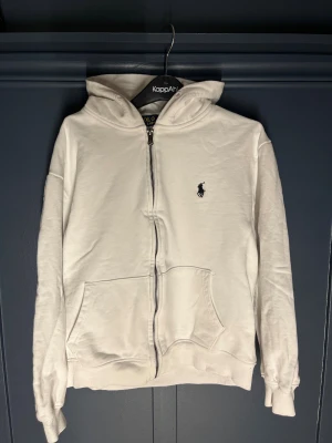 Vit hoodie från Polo Ralph Lauren - Vit hoodie från Polo Ralph Lauren med dragkedja och huva. Klassisk design med broderad logga på bröstet och två stora fickor framtill. och har ribbade muddar vid ärmslut och nederkant. Perfekt för chill dagar. Passar 160cm och neråt. 