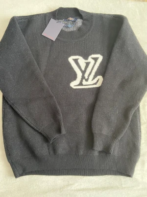 Svarte stickad Louis Vuitton tröja - Svart stickad tröja från Louis Vuitton med stort vitt LV-logo på bröstet. Rund hals med ribbad kant och långa ärmar. Materialet är mjukt och känns lyxigt, perfekt för dig som gillar designerplagg med tydlig branding.
