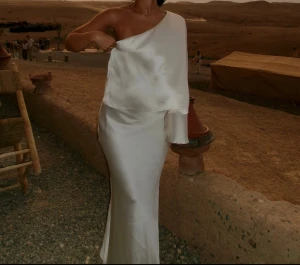 Vit långklänning i satin med oneshoulder - Supersnygg vit långklänning i satin med oneshoulder och elegant draperad ärm. Klänningen har en slät, glansig yta och sitter figurnära över höften. Perfekt för dig som vill sticka ut med en stilren och modern look.