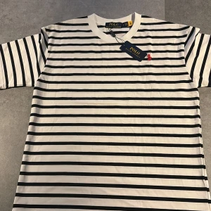 Randig t-shirt från Polo Ralph Lauren - Vit och svart randig t-shirt från Polo Ralph Lauren med klassisk passform och korta ärmar. Tillverkad i mjuk bomull och har den ikoniska röda broderade loggan på bröstet. Perfekt för en clean och stilren look.
