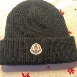 Svart Moncler mössa - Svart ribbstickad mössa från Moncler med klassisk uppvikt kant och broderad logotyp framtill. Mjuk och varm, perfekt för kyliga dagar. Materialet är ull och designen är enkel men stilren, passar dig som gillar exklusiva märken.