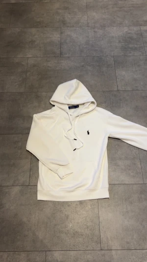 Vit hoodie från Polo Ralph Lauren - Vit hoodie från Polo Ralph Lauren med klassisk svart logga broderad på bröstet. Tröjan har huva med snörning, ribbade muddar och en skön, loose passform. Perfekt för chill dagar och enkel att matcha med jeans eller joggers.
