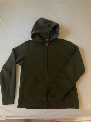 Mörkgrön hoodie från Polo Ralph Lauren - Snygg mörkgrön hoodie från Polo Ralph Lauren med klassisk broderad logga på bröstet. Tröjan har huva, dragkedja framtill och en stor ficka. Tillverkad i mjuk bomull och har långa ärmar med ribbade muddar. Perfekt för chill dagar.