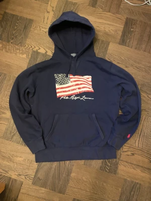 Mörkblå hoodie Polo Ralph Lauren - Säljer en riktigt fet marinblå hoodie från Polo Ralph Lauren med amerikansk flagga tryckt på bröstet och vit signaturtext. Tröjan har huva med snörning, känguruficka och långärmade ribbade muddar. Vid frågor och funderingar, fråga på! 
