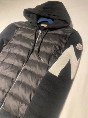 Svart pufferjacka från Mòncler - Svart pufferjacka med stickade ärmar och huva. Framsidan har quiltad polyester och dragkedja, samt två fickor med knapp. Ena ärmen har vit zigzag-mönster och Moncler-logga. Jackan är långärmad och har ribbade muddar.
