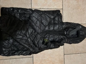 Svart quiltad dunjacka Stone Island - Säljer en svart dunjacka från Stone Island med quiltad design och huva. Jackan har dragkedja framtill och den klassiska Stone Island-loggan på ärmen. Perfekt för dig som vill ha en snygg och varm jacka med streetkänsla.