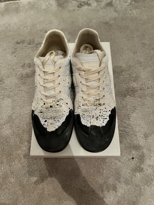 Maison Margiela sneakers med färgstänk - Maison Margiela sneakers i vitt skinn med svart färgstänk och handmålad look. Skorna har beige snörning, gummisula i brunt och en unik design med svart färg över tån. Perfekta för dig som gillar statement pieces och streetstyle. Passar även storlek 41.