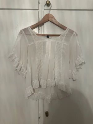 Vit blus med spets och volanger - Säljer en söt vit blus med korta ärmar och romantiska volanger. Blusen har spetsdetaljer längs kanterna och en luftig, flowy passform. Perfekt för dig som gillar en feminin och vintage-inspirerad stil. Fräscha broderade detaljer och rundad halsringning.