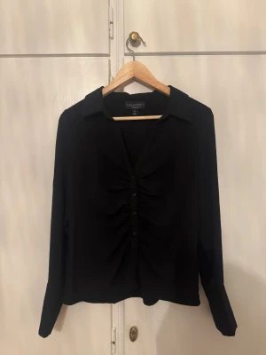 Svart blus med knappar från Laundry - Snygg svart blus från Laundry med krage och knappar framtill. Blusen har ett rynkat parti längs knappraden som ger en cool detalj. Långärmad och i mjukt, lätt material som känns skönt mot huden. Perfekt för dig som gillar stilrena plagg med twist.