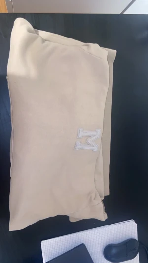 Beige hoodie från Moncler med M-logga - Snygg beige hoodie från Moncler med stor M-logga broderad på bröstet. Tröjan har huva, ribbade muddar och en klassisk känguruficka framtill. Perfekt för dig som gillar stilrena och neutrala färger. Materialet är mjuk bomull och hoodien har en loose passform., liten fläck på luvan 