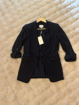 Mörkblå lång kavaj från Vila, strl 34 - Snygg mörkblå lång kavaj från Vila med uppvikta ärmar och klassiska slag. Kavajen har två fickor framtill och är tillverkad i mjuk polyester med lite stretch för extra komfort. Perfekt för en stilren look och enkel att matcha.