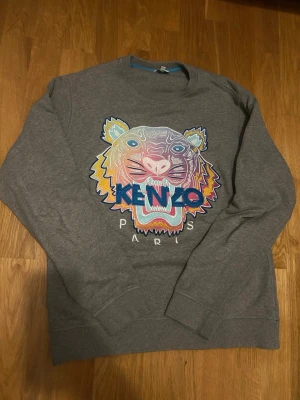 Grå Kenzo sweatshirt med tigertryck - Säljer en grå sweatshirt från Kenzo med ikoniskt broderat tigerhuvud i flera färger och texten 'KENZO PARIS' framtill. 