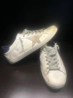 Golden Goose Superstar vita sneakers - Golden Goose Superstar sneakers i vitt skinn med grå mockadetaljer och en glittrig stjärna på sidan. Skorna har blå hälkappa, grå snörning och svart sula med fiskbensmönster soppa. Klassisk rund tå och låg profil. Märkeslogga på sidan och häl.