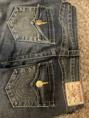 True Religion Joey Low Rise Flare Jeans - Säljer ett par mörkblå True Religion Joey jeans med låg midja och flare-ben. Klassiska kontrastsömmar, snygga fickdetaljer med guldknappar och ikonisk logga bak. Tillverkade i slitstarkt jeansmaterial. Endast använt fåtal gånger ( nypris 1500 kr)
