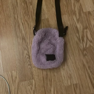 Lila fluffig axelväska - Supersöt lila axelväska med fluffig framsida och svart justerbar axelrem. Baksidan är i slätt tyg och väskan har en liten svart patch framtill. Perfekt storlek för mobilen och småprylar. Har en liten fläck som är från ett läppglans men det är ingen som syns eftersom den är på insidan av väskan, annars supersöt lila axelväska💜