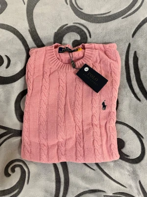 Rosa kabelstickad tröja Polo Ralph Lauren - Säljer en rosa kabelstickad tröja från Polo Ralph Lauren. Tröjan har rund hals, långa ärmar och den klassiska broderade loggan på bröstet. Perfekt för dig som vill ha en stilren och tidlös look med en mjuk känsla.