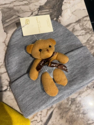 Grå mössa med nalle från Poyraz Sapka . Barn - Supersöt grå mössa med en mjuk liten nalle fastsydd framtill. Nallen har ett brunt band med texten 'Love' runt halsen. Mössan är ribbstickad och har en uppvikt kant för extra stilpoäng. Perfekt accessoar för dig som vill sticka ut lite extra.