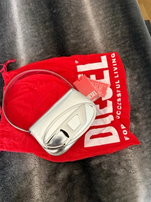 Silver axelväska från Diesel - Säljer en futuristisk silverfärgad axelväska från Diesel med coolt 3D-mönster på framsidan och magnetlås. Väskan har ett huvudfack och en innerficka, samt matchande axelrem. Längre axelband medföljer! 