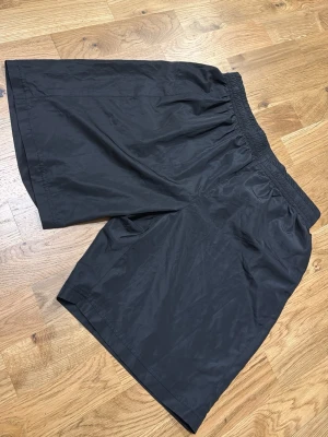 Svarta loose shorts i syntet - Säljer ett par svarta, loose-fit shorts med elastisk midja. Tillverkade i ett lätt och glansigt syntetmaterial som känns svalt och skönt. Perfekta för sommardagar eller när du vill ha något bekvämt och luftigt.