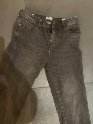 Mörkgrå jeans från Jack & Jones, 146 - Säljer ett par mörkgrå jeans från Jack & Jones i storlek 146. Klassisk femficksmodell med raka ben och snygga sömmar på bakfickorna. Jeansen har dragkedja och knapp framtill och är tillverkade i ett mjukt bomullsmaterial med lite stretch.