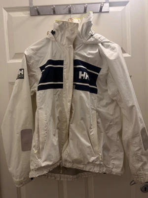 Vit vindjacka från Helly Hansen - Säljer en vit vindjacka från Helly Hansen med marinblåa ränder och logga på bröstet och ärmen. Jackan har hög krage, huva och dragkedja framtill. Tillverkad i polyester och har långa ärmar med reflexdetaljer. Perfekt för blåsiga dagar!