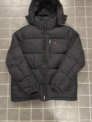 Svart pufferjacka  - Säljer en svart pufferjacka  med huva och dragkedja framtill. Jackan har en röd broderad logga på bröstet, flera fickor med dragkedja och quiltad design. Perfekt för kalla dagar och riktigt snygg till streetwear-stilen. Finns i storlek S och M 