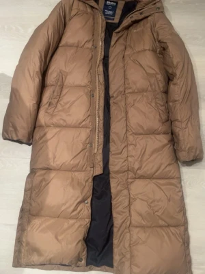 Lång brun dunjacka från Svea XS - Lång, brun dunjacka från Svea i storlek XS, men passar S. Jackan har huva, dragkedja och knappar framtill.Perfekt för kalla dagar och har en clean look med diskret Svea-logga på bröstet.