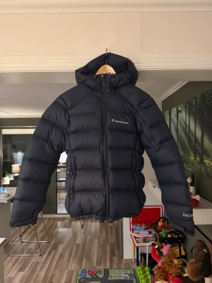 Mörkblå dunjacka från Peak Performance - Säljer en mörkblå dunjacka från Peak Performance med huva och dragkedja framtill. Jackan är vadderad och har en klassisk pufferstil som håller dig varm under kalla dagar. Perfekt för dig som vill ha en snygg och funktionell vinterjacka. Har använt den som stalljacka men den är vädrad och tvättad, ingen hästlukt vad jag känner. Priset kan diskuteras 