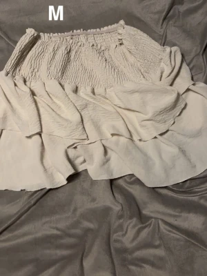 Vit volangkjol med smock, stl M - Supersöt vit kjol med bred smock i midjan och dubbla volanger. Kjolen har en luftig och flowy känsla, perfekt för varma dagar. Passar dig som gillar en feminin och avslappnad stil.