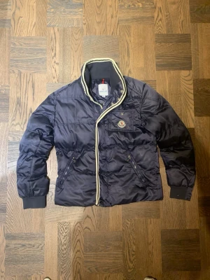 Mörkblå Moncler pufferjacka - Säljer en mörkblå pufferjacka från Moncler. Vet ej om jackan är äkta eller inte då den hängt i garderoben väldigt länge där av ett rimligt pris. Jackan är i väldigt bra skick men saknar luva och en del av dragskedjan den man håller i är lite trasig men det är fortfarande lätt att stänga och öppna. Vid frågor eller funderingar fråga på! 