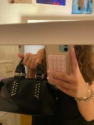 Svart handväska med nitar - Säljer en svart handväska i skinnimitation med coola silverfärgade nitar på handtagen. Väskan har dubbla handtag, silverfärgade metalldetaljer och dragkedja upptill. Perfekt storlek för det viktigaste och ger en edgy vibe till din outfit.