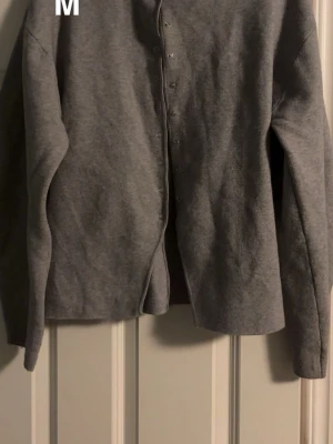 Grå kofta med knappar, storlek M - Mysig grå kofta i mjukt material med långa ärmar och knappar framtill. Enkel och clean design som passar perfekt till lager på lager under höst och vinter. Snygg att matcha med jeans eller kjol.