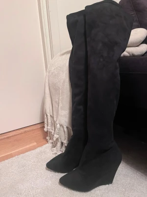Svarta overknee stövletter i mocka - Svarta overknee stövletter med kilklack (10cm) och spetsig tå. Skorna är tillverkade i mjuk mocka och har en elegant, hög siluett som går över knät.  Storlek 37. Mycket bra skick! Säljs på flera sidor, först till kvarn. 